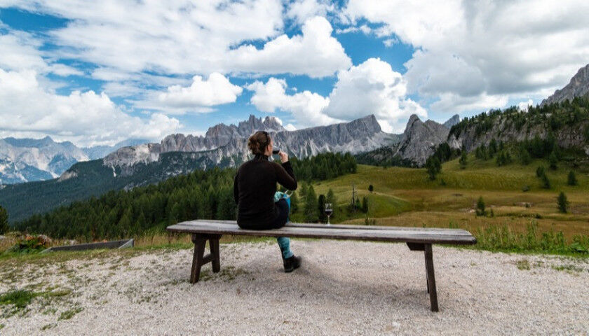 young woman sitting at bench, drinking glass of red wine, looking at Cima Ambrizzola in front of Rifugio Cinque Torri. Dolomites_20221010
